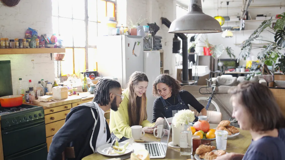 Huisgenoten zitten samen aan de keukentafel met koffie, fruit en een laptop
